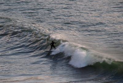 [An all-black (because of wet-suit including a hood and gloves) figure surfs about a 5 foot wave which is collapsing to his left but still forming to his right.]